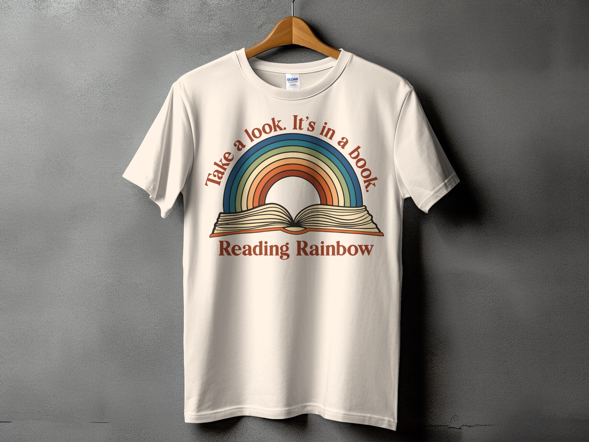 Cream t-shirt with colorful rainbow and book graphic, text reads "Take a look. It's in a book Reading Rainbow"