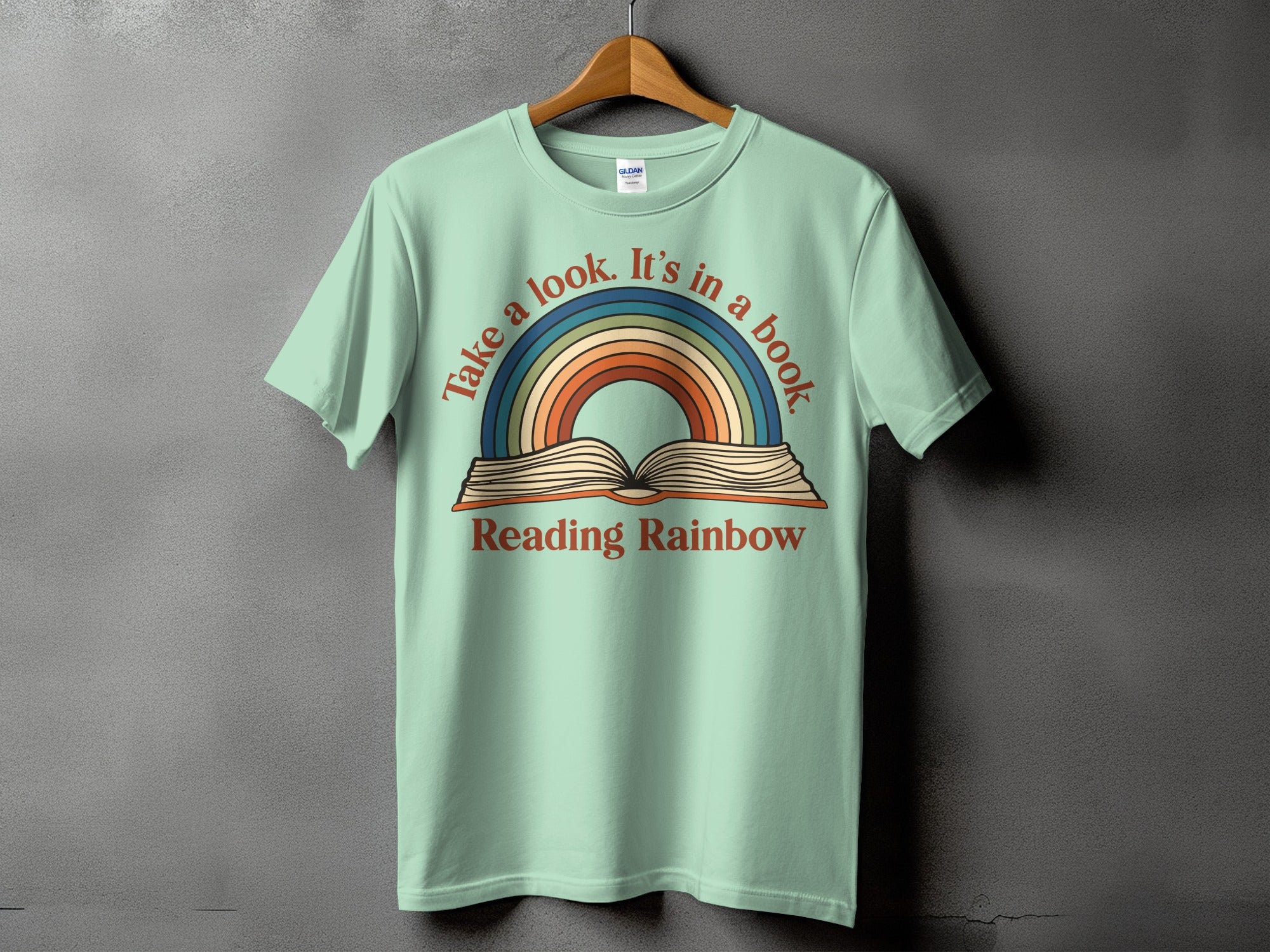 Light green t-shirt with open book and rainbow graphic, text reads "Take a look. It's in a book. Reading Rainbow."