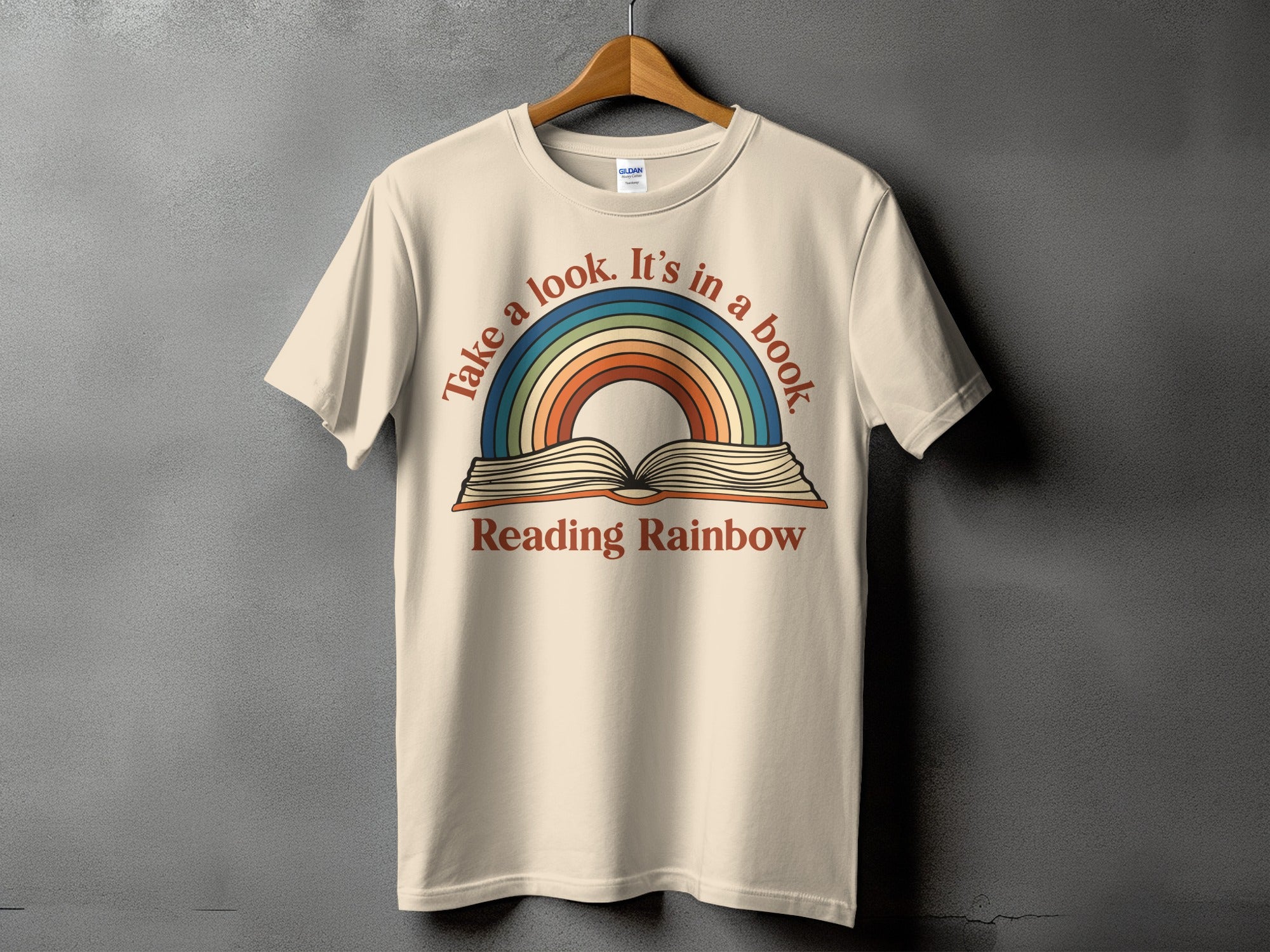 T-shirt with open book and rainbow graphic, text reads "Take a look. It's in a book. Reading Rainbow."
