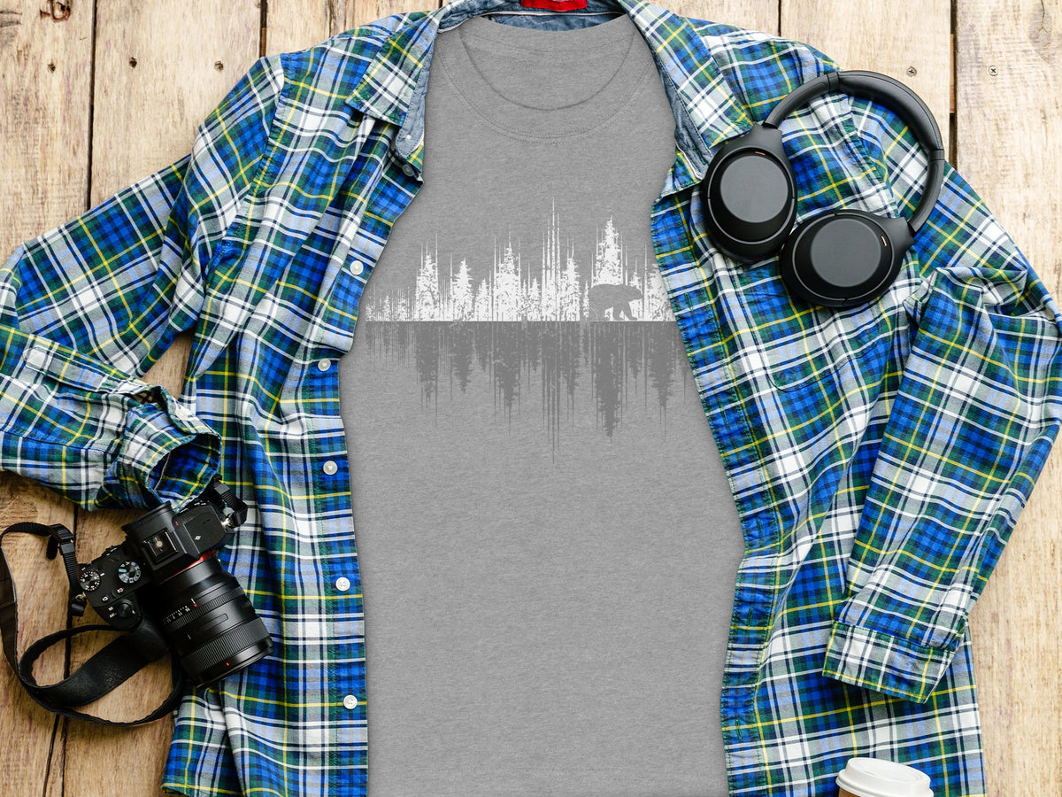 Gray t-shirt with white graphic of trees and a walking bear, laid flat with plaid shirt, camera, and headphones.