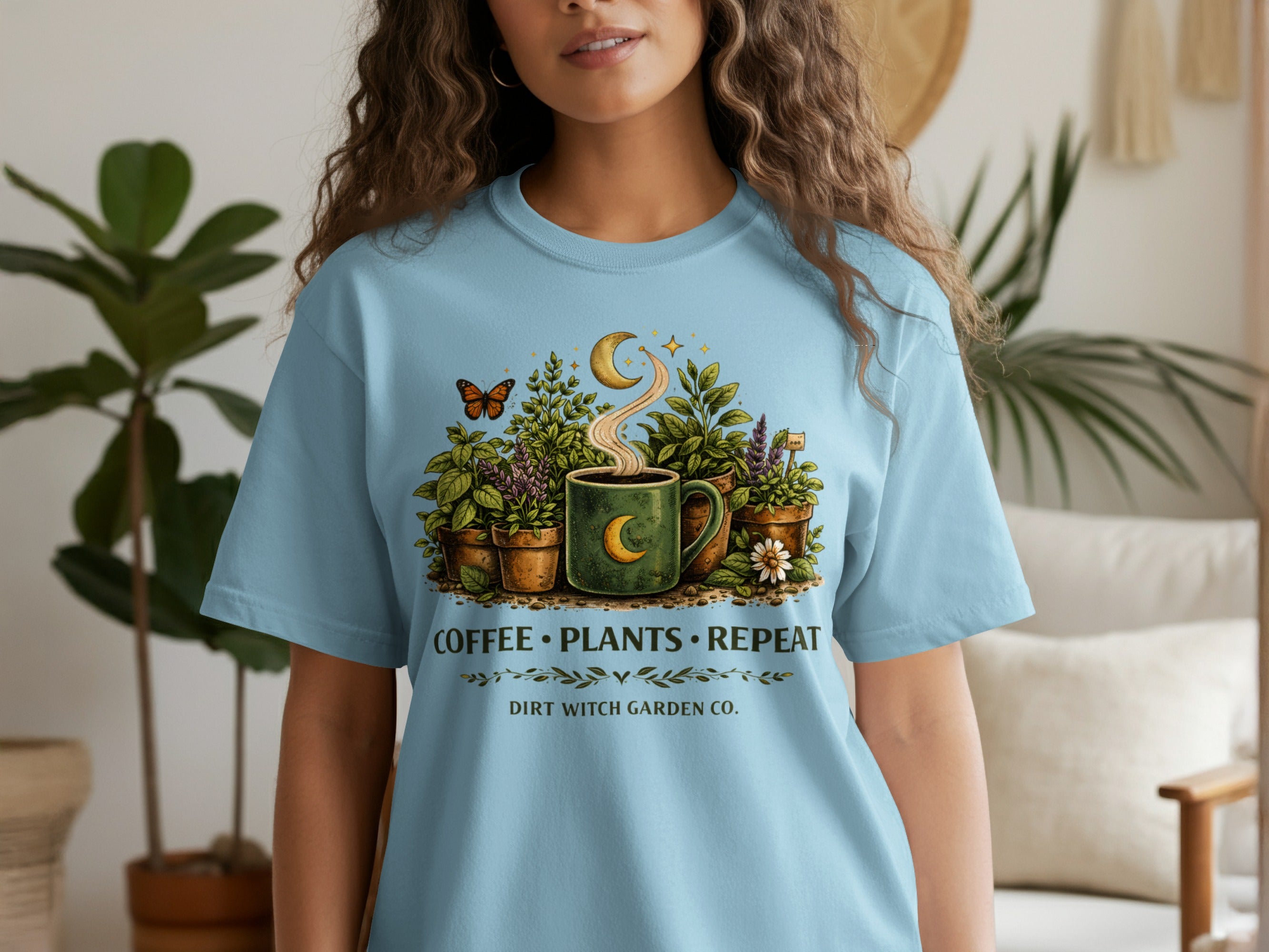 Light blue t-shirt with graphic of plants, coffee cup, moon, and butterfly. Text reads "Coffee Plants Repeat. Dirt Witch Garden Co."