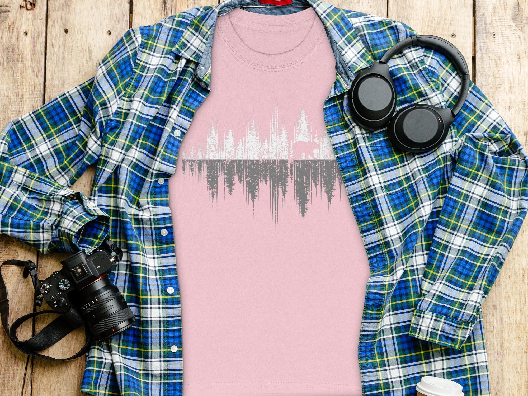 Pink t-shirt with forest and bear graphic, plaid shirt, headphones, and camera.
