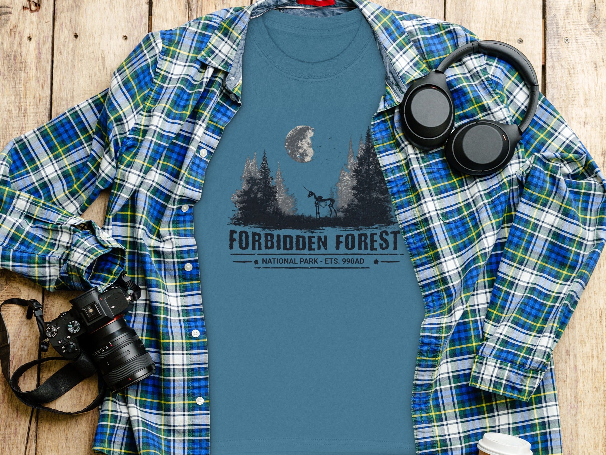 T-shirt with "Forbidden Forest National Park" graphic, laid with a plaid shirt, headphones, and camera.