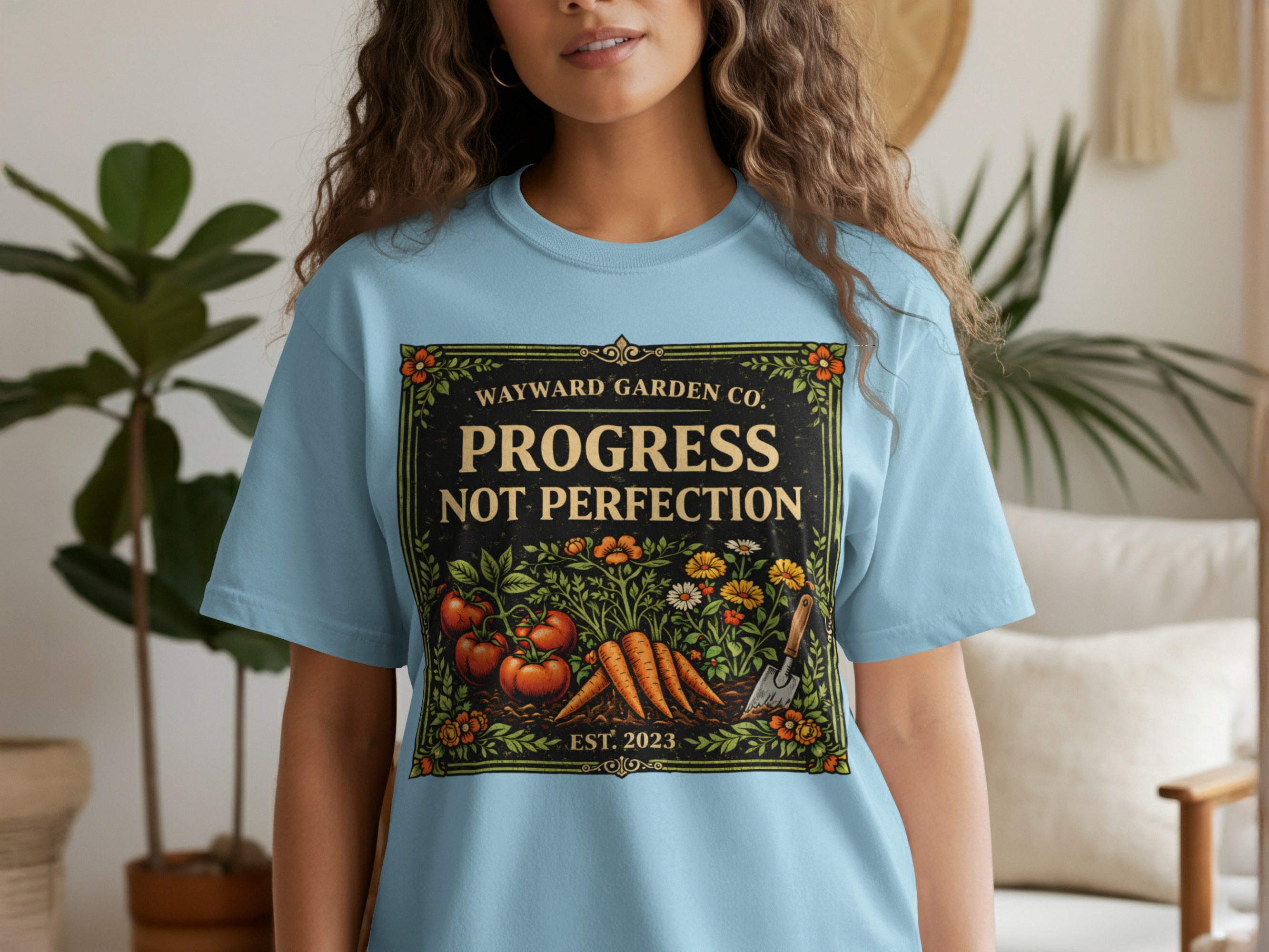 Light blue t-shirt with "WAYWARD GARDEN CO. PROGRESS NOT PERFECTION EST. 2023" text and garden illustration.