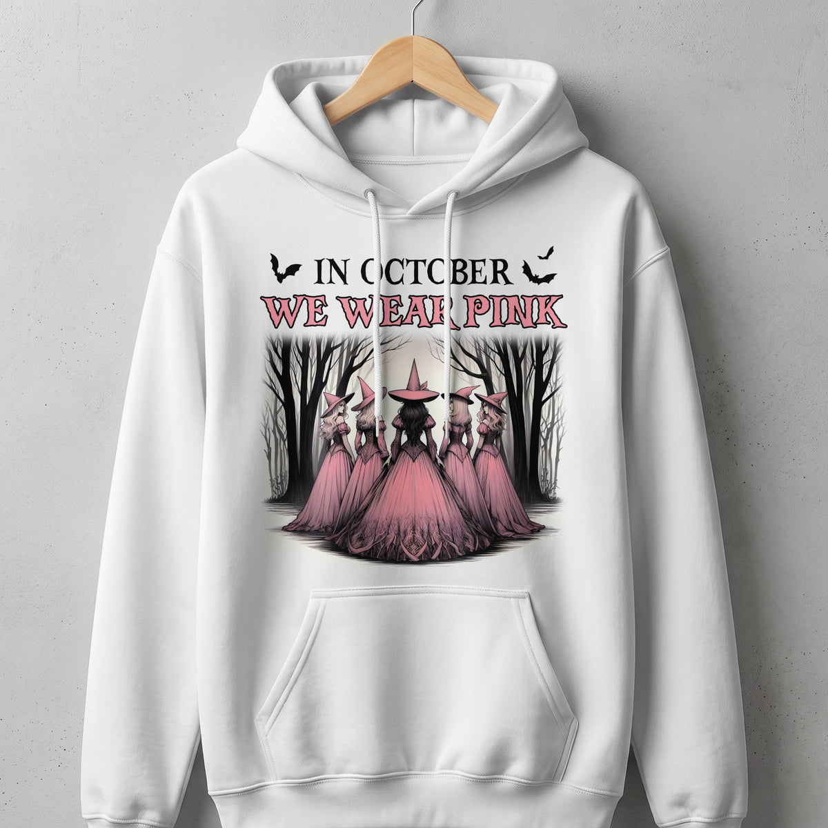 White hoodie with "In October We Wear Pink" text and witches in a forest.