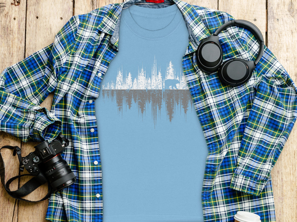 Light blue t-shirt with white and gray forest and bear graphic, over plaid shirt.