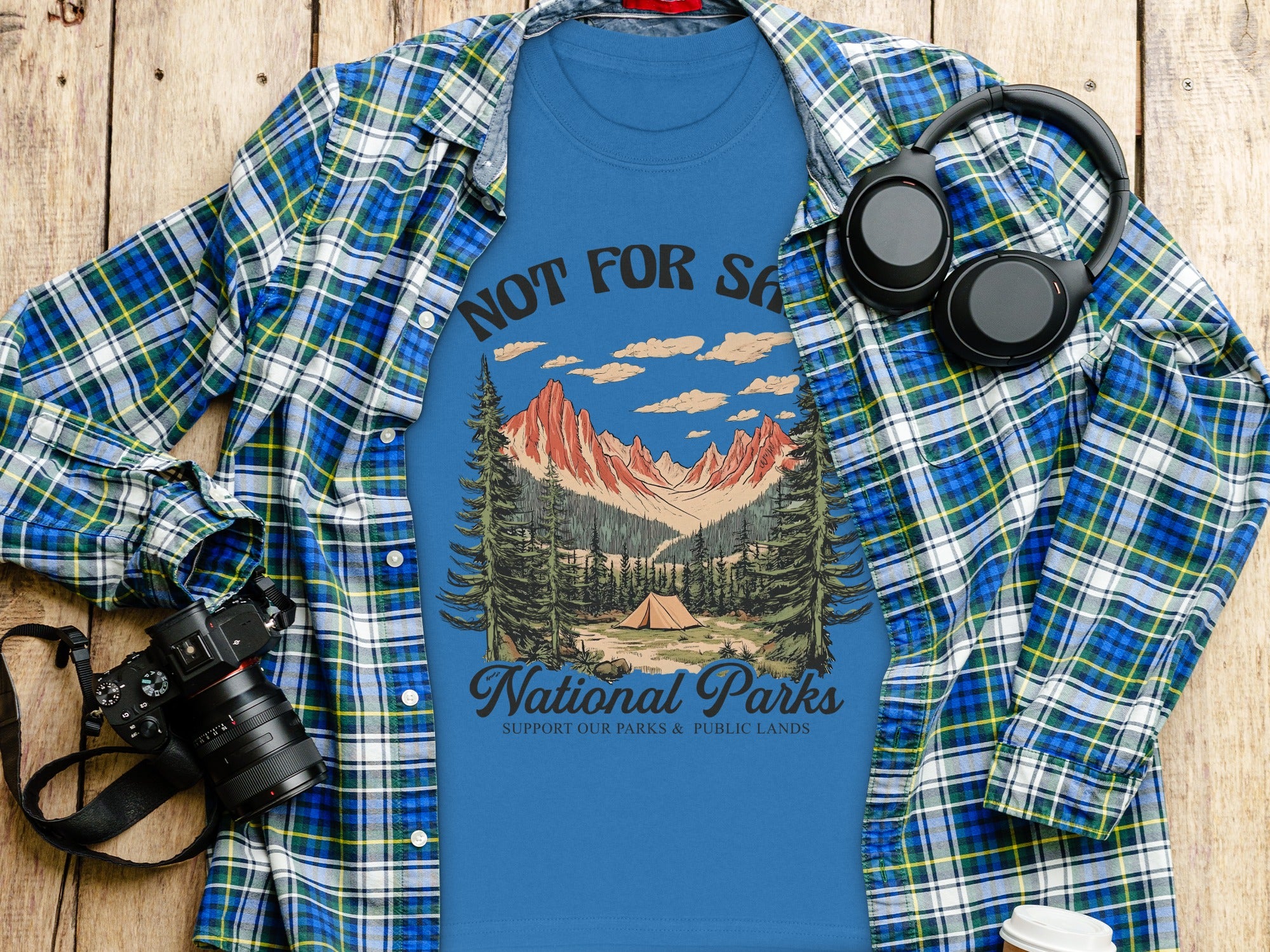 Blue t-shirt with National Parks graphic, worn open over plaid shirt, with headphones and camera.