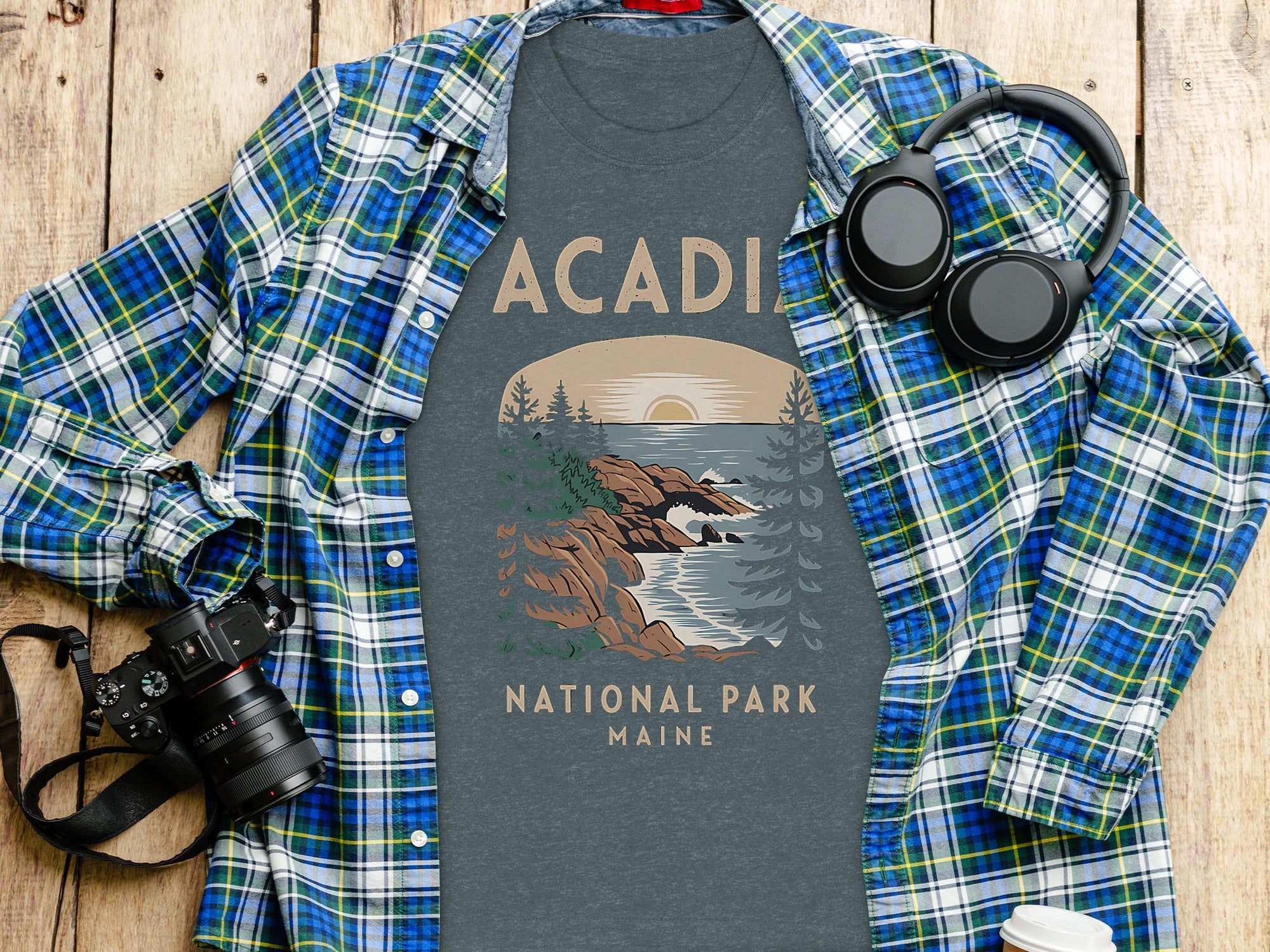 T-shirt with "Acadia National Park Maine" text and scenic coastal artwork, laid flat with a plaid shirt, camera, and headphones.