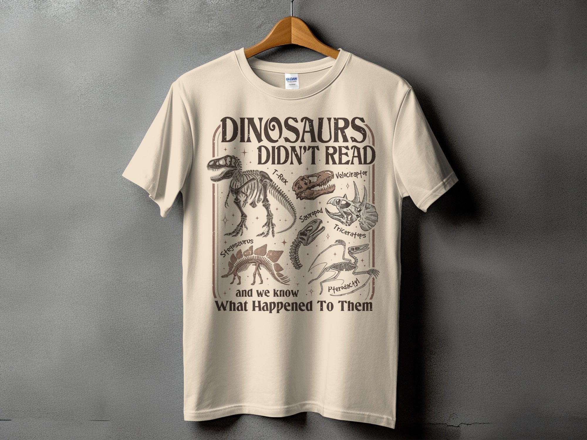 Cream t-shirt with a graphic of dinosaur skeletons and "Dinosaurs Didn't Read" text.