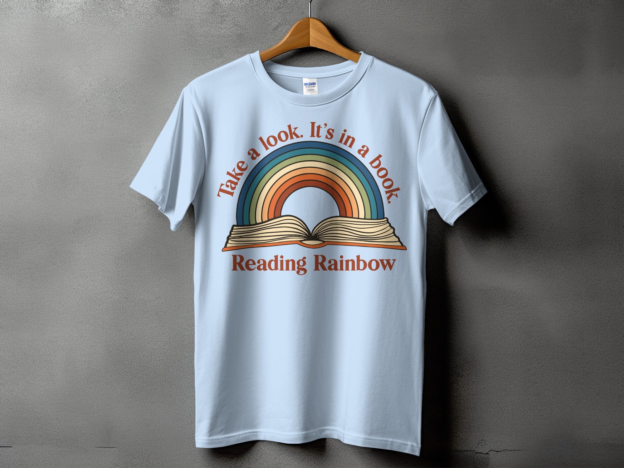 Light blue t-shirt with a rainbow arching over an open book, text reads "Take a look. It's in a book. Reading Rainbow".