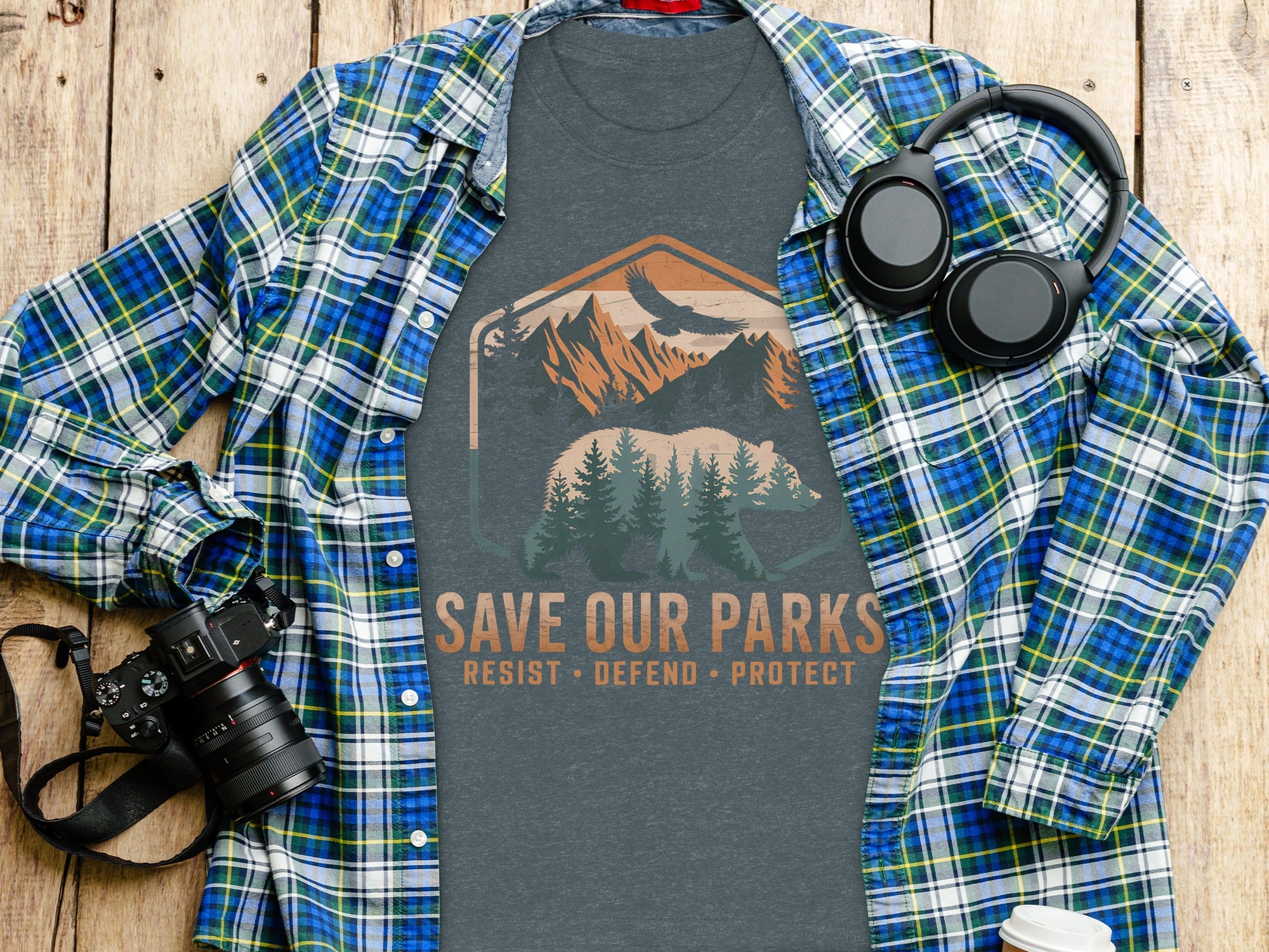 T-shirt with "Save Our Parks" bear and mountain graphic, flanked by plaid shirt, headphones, and camera.