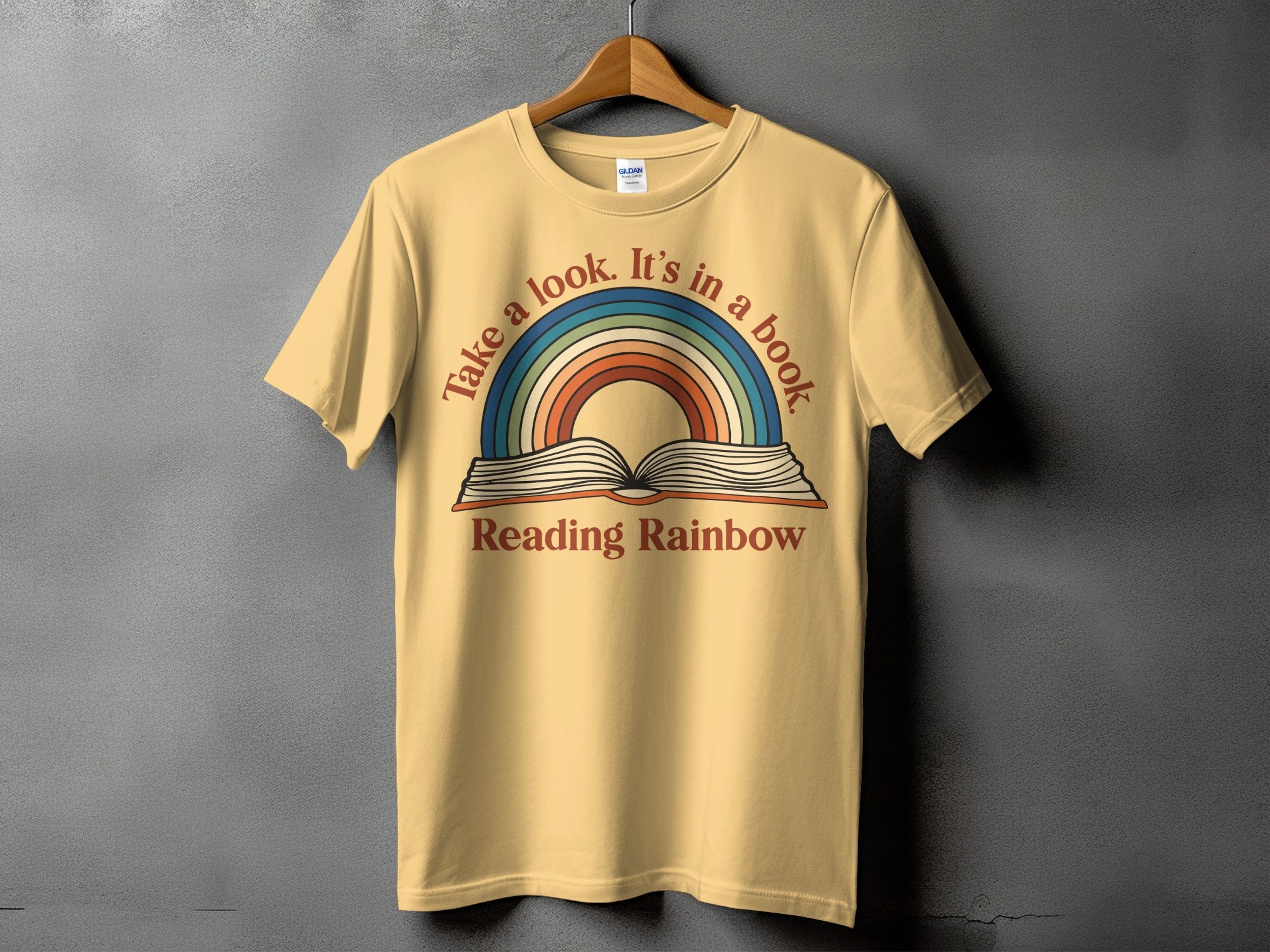 Yellow t-shirt with a graphic featuring an open book and rainbow, text reads "Take a look. It's in a book."