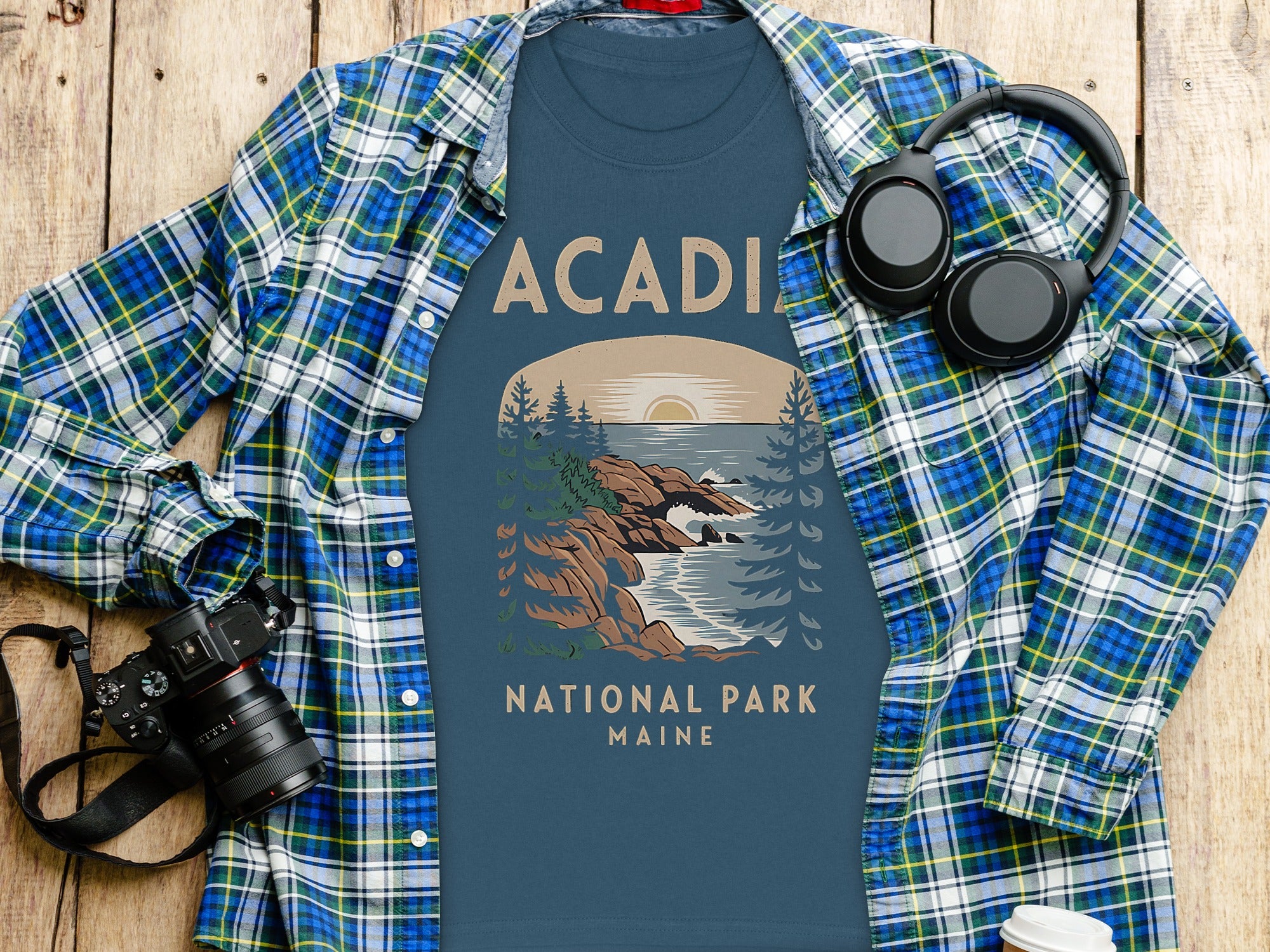 Blue t-shirt with Acadia National Park Maine graphic, shown with plaid shirt, headphones, and camera.