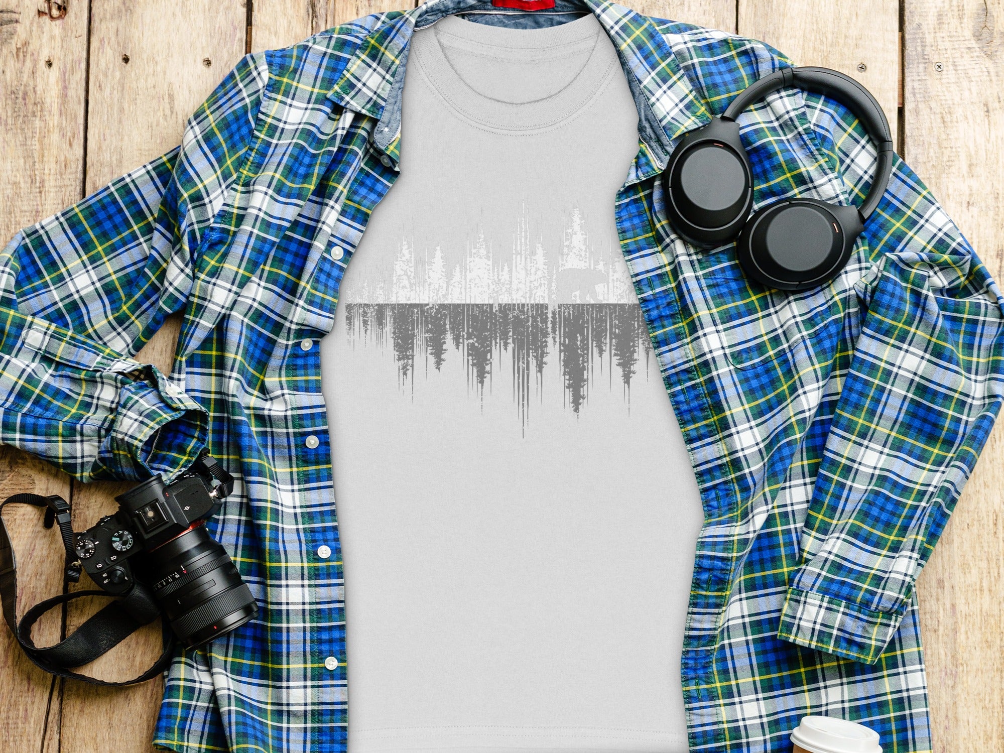 Grey t-shirt with forest and bear graphic, framed by plaid shirt, headphones, and camera.