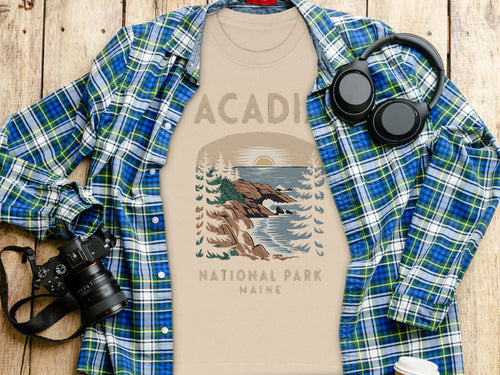 Light tan t-shirt with Acadia National Park Maine graphic, next to a plaid shirt and camera.