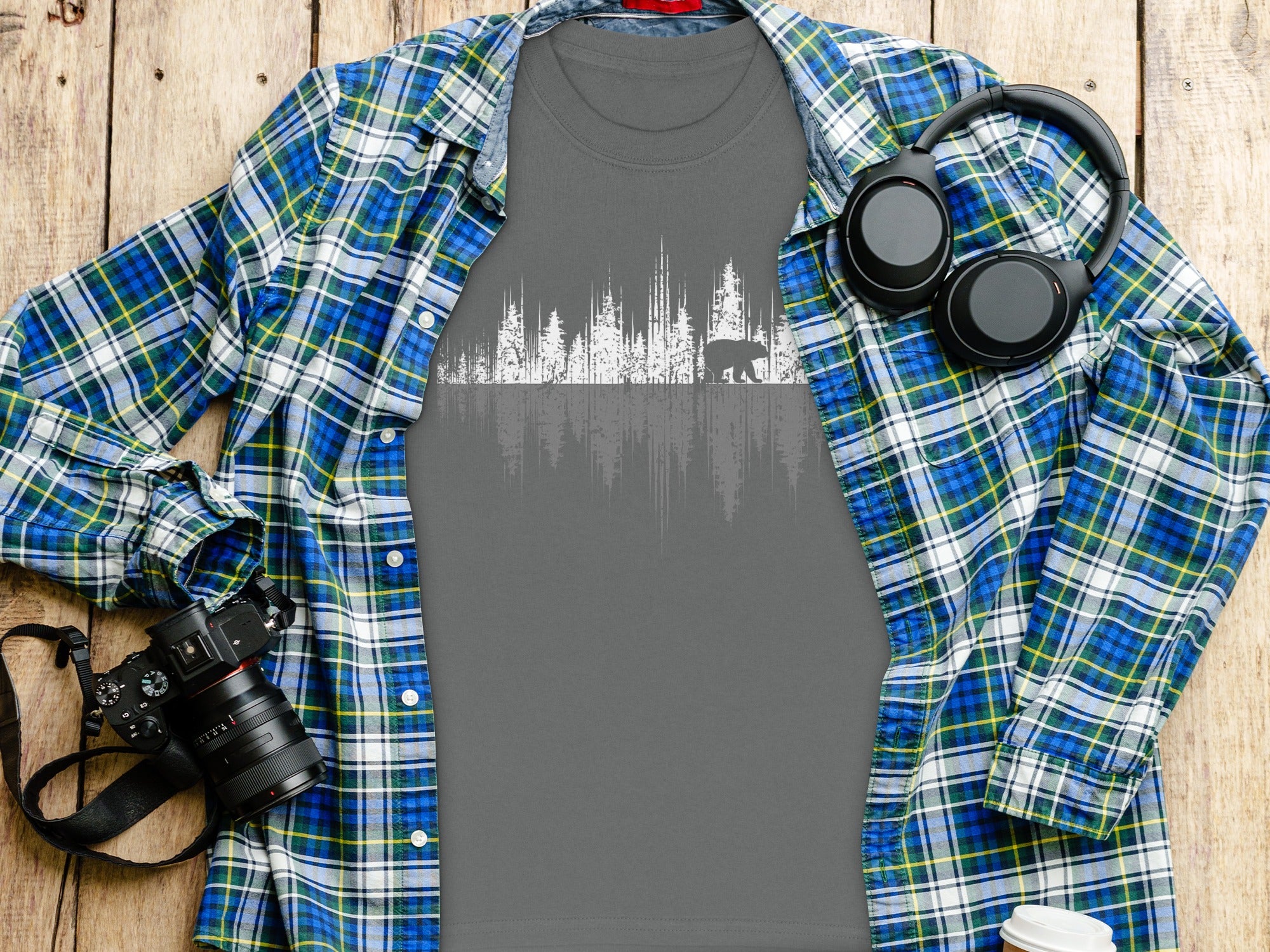 Gray t-shirt with white graphic of bear and trees, next to flannel shirt, camera, and headphones.